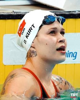 Yüzme havuzunda kenarda duran yüzücü Defne Kurt. Üzerinde beyaz bir bone var ve bonede Türk bayrağı ile "TUR" ve "D. KURT" yazıları yer alıyor. Yüzünde ve omzunda dövmeler bulunuyor. Gözlüğü bonenin üstünde duruyor. Başını hafifçe yukarı kaldırmış ve suda dinleniyor. Arka planda sarı bir zemin üzerinde beyaz bir tabela var, "TRT SPOR" logosu sağ altta yer alıyor.