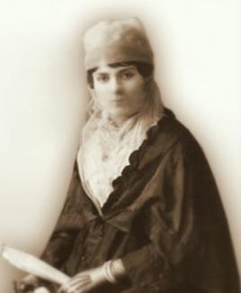 Mihrî Hatun’un başında bir örtü/bere ve boynunda bir şal var. Üzerinde koyu renk, uzun kollu geleneksel bir elbise bulunuyor. Hafifçe bulanık bir şekilde poz vermiş ve elinde bir şey tutuyor. Zemin açık renk ve arka plan sade.