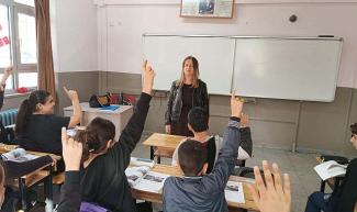 Bir sınıf ortamı. Sınıfın önünde, beyaz tahtanın önünde bir kadın öğretmen ayakta duruyor. Karşısında sıralarda oturan öğrenciler var. Birkaç öğrenci elini kaldırmış, muhtemelen öğretmenden söz hakkı almak istiyorlar ya da soruya cevap vermek istiyorlar. Sıralarda açık kitaplar ve defterler var. Pencere kenarında Türk bayrakları dikkat çekiyor. Sınıfın üst köşesinde Atatürk’ün resmi ve bir yazı asılı. Öğrencilerin çoğu öğretmene odaklanmış durumda.