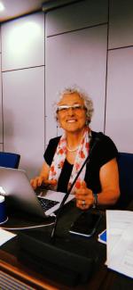 Nazik Işık, kıvırcık beyaz saçlı, gözlüklü ve gülümsüyor. Bir bilgisayarın önünde oturuyor ve kulaklık takıyor. Boynunda inci bir kolye ve üzerindeki siyah kıyafetin üstüne kırmızı çiçek desenli açık renkli bir fular takmış. Elleriyle bilgisayarın klavyesine dokunuyor. Önünde bir mikrofon, masada ayrıca bir telefon, mavi bir kupa ve çeşitli kağıtlar bulunuyor. Mutlu görünüyor. Bir toplantı odasına benzer modern bir mekânda oturuyor.