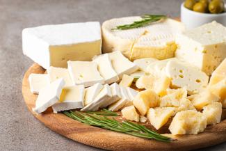 Ahşap bir servis tahtası üzerinde çeşitli peynirler. Ön planda krem rengi kabuklu, içi sarımsı beyaz olan brie veya camembert peyniri dilimlenmiş halde. Yanında sarımsı, iri parçalara ayrılmış parmesan peyniri. Arkada yuvarlak formda ve açık renkli bir peynir ile delikli bir beyaz peynir bloğu. Tahtanın üzerinde ve peynirlerin arasında taze biberiye dalları. Arka tarafta küçük bir kapta yeşil zeytinler görünüyor.