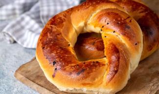 Açık kahverengi ve yuvarlak biçimli bir çörek. Üzeri hafifçe kızarmış ve bazı yerlerinde çörek otu serpilmiş. Yuvarlak şekli ve parlak kabuğuyla taze görünüyor. Çöreğin arka tarafında bir tane daha var. Çörek tahtası üstünde ve arka planda gri-beyaz çizgili bir bez yer alıyor. Çörek çok yumuşak ve kabarık gözüküyor.