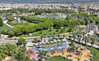Şehir parkı ve çocuk oyun alanı. Ön planda, çeşitli oyun aletlerinin bulunduğu renkli ve geniş bir çocuk oyun alanı var. Salıncaklar, kaydıraklar ve tırmanma alanları görülebiliyor. Etrafı palmiye ağaçlarıyla çevrili. Parkta yürüyüş yolları, bolca çimenlik alan ve gölgelik alanlar mevcut. Daha geride ağaçlarla kaplı geniş bir park var. Parkın hemen ilerisinde şehir merkezine ait binalar ve apartmanlar sıralanmış; arka planda dağlar gözüküyor. Gökyüzü açık ve hava güneşli görünüyor. Her tarafta yeşil alan ve