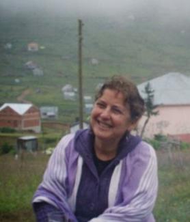 Açık alanda çekilmiş bir fotoğraf. Ön planda, açık mor renkli bir hırka giymiş ve gülümseyen bir kadın var. Arka planda sisli bir hava hâkim; bir tepenin yamacında dağınık şekilde serpiştirilmiş, çatıları metal ya da kiremitten birkaç ev ve köy manzarası bulunuyor. Çimenler ve yeşil alan, ortamın doğal olduğunu gösteriyor.