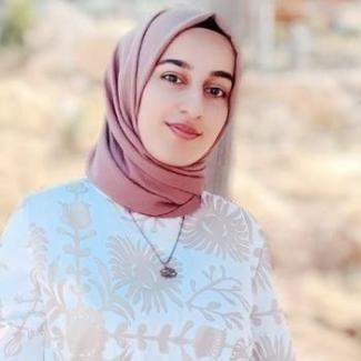 Açık ortamda çekilmiş bir fotoğraf. Açık tenli, pembe tonlarında başörtüsü takan bir kadın. Üzerinde beyaz ve açık kahverengi desenli bir bluz var. Boynunda kolye takıyor. Arka planda doğa ve bulanık ağaçlar var. Kadın hafif bir tebessümle kameraya bakıyor.