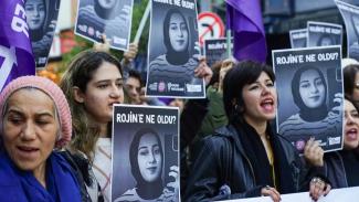Kadınlardan oluşan bir grup, sokakta topluca protesto yapıyor. Ellerinde bir genç kadının başörtülü fotoğrafının bulunduğu afişler tutuyorlar. Afişlerin üstünde büyük harflerle "ROJİN'E NE OLDU?" yazıyor. Gruptakilerin yüz ifadeleri ciddi ve bağırıyorlar, slogan atıyorlar. Bazı kişiler mor renkli bayraklar taşıyor.