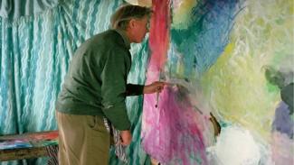 Resim yapan bir adam. Ortamda mavi-beyaz dalga desenli bir perde var. Adam yeşil bir kazak ve kahverengi pantolon giymiş, sol profilinden görünüyor. Önünde büyük bir tuval var ve renkli, soyut bir tablo üzerinde çalışıyor; tabloda mavi, pembe, sarı gibi canlı renkler hâkim. Adam elinde bir fırça tutuyor ve tabloya odaklanmış resim yapıyor. Arkada bir tahta masa var, üzerinde boyalar veya sanat malzemeleri bulunuyor.