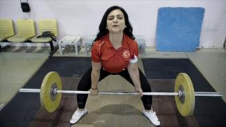 Ağırlık kaldıran Gülistan Özdemir, kırmızı milli takım tişörtü giymiş ve üzerinde Türk bayrağı simgesi var. Yerde bulunan halter çubuğunu iki eliyle kavrayıp kaldırmak üzere pozisyon almış. Arka planda sarı sandalyeler, siyah bir çanta ve beyaz bir sandalye ile mavi bir minder görünüyor.