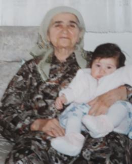 Koltukta oturan yaşlı bir kadın ve kucağında bir bebek. Kadın başında açık renkli bir eşarp ve desenli koyu renkli bir elbise giymiş. Bebek açık renkli kıyafetler ve beyaz patik giymiş. Arka planda açık renk bir duvar ve koltuk var. Kadın hafif gülümsüyor ve bebek sakince oturuyor.