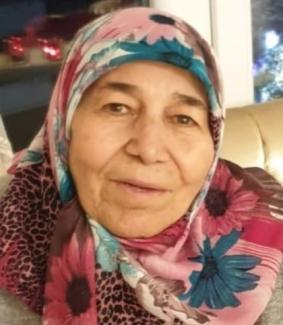 Şükriye Hanım Bir koltukta oturuyor. Başörtüsü pembe ve mavi ağırlıklı, üzerinde büyük çiçek desenleri var. Hafifçe gülümsüyor ve doğrudan kameraya bakıyor. Arka planda bir pencere ve silik şekilde parlak ışıklar görünüyor.