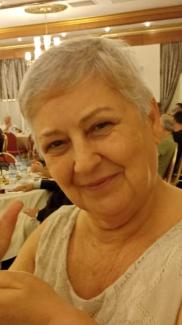 Yakın çekim fotoğraf. Kısa, gri saçları var ve hafif gülümseyerek kameraya bakıyor. Üzerinde krem veya açık renkli, dantel desenli bir bluz bulunuyor. Arka planda bir restoran ya da davet salonu gibi bir ortam görünüyor; masada su şişeleri duruyor ve diğer insanlarla dolu yuvarlak masalar bulunuyor. Ortamda sıcak bir ışık hakim.