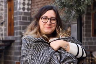 Diyarbakır'da yer alan eski bir Zerzevan Konağı'nın koyu gri taş duvarlarıyla kaplı avlusunda, bir zeytin ağacının altında kucağında tekir bir kedi ile muzip şekilde kameraya gülümsüyor. Saçları dağınık ve kediyi omuzlarına aldığı kalın şalla bebek gibi sarmış. Kedi bu durumdan biraz mutsuz görünüyor.