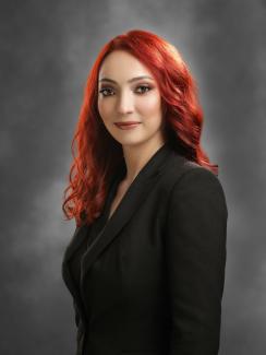 Alev kırmızısı dalgalı, uzun saçları, siyah ceketiyle ayakta gülümsüyor.