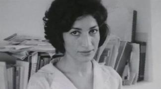 Kısa, siyah, dalgalı saçları, beyaz bluzuyla kitaplarla dolu bir rafın önünde ciddiyetle bakıyor. (Siyah-beyaz fotoğraf)