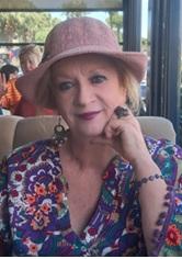 Açık renkli şapka takmış, kısa sarı saçlı, renkli, çiçek desenli bir bluz giymiş. Kolunda mor boncuklu bir bileklik, büyük bir yüzük ve sarkık küpeleri var. Yüzünde hafif bir tebessümle kameraya bakıyor ve bir elini çenesine yaslamış. Arka planda başka insanlar ve dış mekân bir kafe veya restoran atmosferi görünüyor.