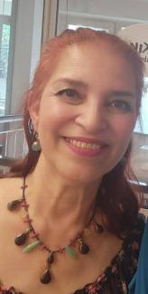 Uzun, kızıl-kahverengi saçlı ve saçları arkaya doğru toplanmış. Yüzünde hafif bir makyaj bulunuyor; gözlerinde eyeliner ve pembe tonlarında bir ruj sürülmüş. Gülümsüyor. Üzerinde çiçek desenli bir bluz ve boynunda renkli taşlardan oluşan gösterişli bir kolye var. Sol kulağında sarkık bir küpe takılı. Arka planda cam bir pencere ve dışarıda ağaçlar görünüyor.