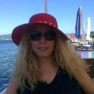 Deniz kıyısındaki bir mekânda, kırmızı melon şapkası, koyu renk güneş gözlükleri ve sarı uzun saçları, kolsuz siyah bluzu; ince bir kolyesi var.