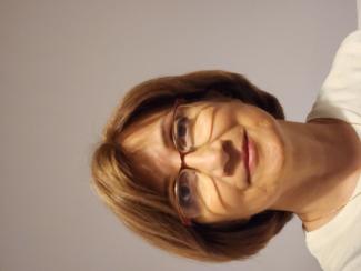 Kısa açık kahverengi saçlı, gözlük takan, açık tenli Nalan Yılmaz duvar önünde duruyor. Beyaz bir üst giymiş ve hafifçe gülümsüyor. Arka plan sade, açık renkli ve herhangi bir detay yok. Yılmaz doğrudan kameraya bakıyor.