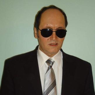 Koyu renk ceket içinde beyaz gömlek ve enine siyah çizgili gri kravatı, koyu renk güneş gözlüğüyle ağzı hafif aralık, ciddi bir duruş sergiliyor.