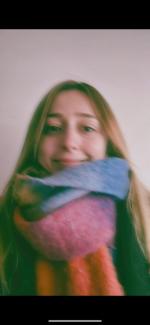 Zeynep Selcen Toprak’ın yüzünde hafif bir gülümseme var. Saçları açık kahverengi ve omuzlarına dökülüyor. Boynuna renkli bir atkı dolanmış, atkıda mavi, gri, pembe ve turuncu gibi canlı renkler var. 