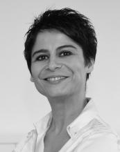 Siyah saçları, beyaz bluzu var, kendine güvenle gülümsüyor.