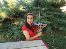 Yeşil bir alanda bir piknik masasında oturmuş, sol elinde kemanı sağ elinde yayı, kırmızı elbisesiyle ve siyah uzun saçlarına taktığı tacıyla ,gülümsüyor.