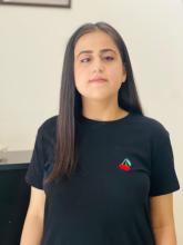 Düz beyaz arka plan önünde duran Berfin Geldi, uzun, düz ve koyu renkli saçlı. Siyah tişört giymiş ve tişörtün sol göğüs kısmında küçük, kırmızı bir kiraz deseni bulunuyor. Doğrudan kameraya bakıyor, yüzünde hafif bir gülümseme var. Arka planda sol üst köşede çerçeveli bir tablo veya fotoğrafın köşesi görünüyor.