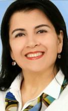 Gülen bir portre. Koyu renk, düz ve omuz hizasında saçları var. Kırmızı ruj sürmüş ve hafif bir makyaj yapmış. Küçük yuvarlak küpeleri dikkat çekiyor. Üzerinde beyaz bir gömlek ve renkli, mavi ağırlıklı bir fular var. Arka plan tek renk, açık mavi.