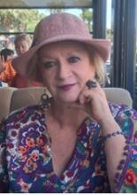 Açık renkli şapka takmış, kısa sarı saçlı, renkli, çiçek desenli bir bluz giymiş. Kolunda mor boncuklu bir bileklik, büyük bir yüzük ve sarkık küpeleri var. Yüzünde hafif bir tebessümle kameraya bakıyor ve bir elini çenesine yaslamış. Arka planda başka insanlar ve dış mekân bir kafe veya restoran atmosferi görünüyor.