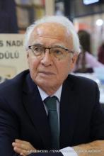 Kısa beyaz saçlı ve beyaz kaşlı. Gözlük takıyor ve ciddi bir ifadeyle ileriye bakıyor. Üzerinde koyu renk bir takım elbise, beyaz gömlek ve yeşil bir kravat var. Arka planda bulanık bir şekilde başka insanlar ve kitap fuarına ait bir ortam görünüyor. Fotoğrafın alt kısmında “KADİR İNCESU / TÜYAP İSTANBUL KİTAP FUARI 202” yazısı yer alıyor.