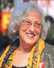 Beyaz, dalgalı, küt saçları; çerçevesiz gözlükleri ile başını soluna çevirmiş, kocaman gülümsüyor. Haki yapraklı bluzunun üzerinde sarı ağırlıklı desenli şalı ve kolyesi var.