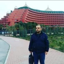 Kırmızı renkli, büyük ve dikkat çekici mimarisiyle öne çıkan bir otel binasının önünde, kaldırımda duran Mehmet Yıldırım, koyu renkli bir mont ve pantolon giymiş, sağ elinde siyah bir çanta tutuyor. Arka planda otelin kubbeli çatısı ve kat kat balkonları görünüyor. Yanında tel örgülü bir çit ve yeşillikler var.