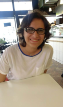 Kısa koyu kahverengi saçları ve koyu çerçeveli gözlüğüyle gülümsüyor. Yakasında incecik mavi biyesi olan beyaz bluzu var.